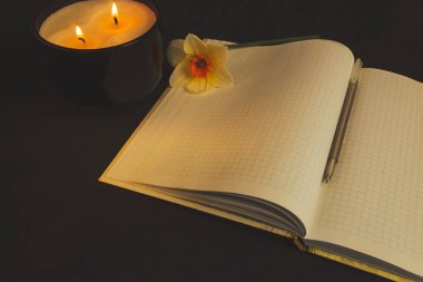 Kalemli defter, sarı narsisli çiçeğin yanında ve koyu arkaplanda yanan bir mum..