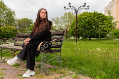 Bir manken şehir parkında bir bankta oturur ve başka yere bakar. Siyah elbiseli, beyaz spor ayakkabılı ve siyah saçlı bir kız. Ciddiyet ve zarafet kavramı. Açık havada bahar fotoğrafı..