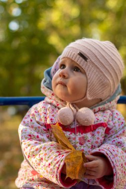 Bir çocuk dışarıda oturur, bir ağaçtan yaprak tutar ve başka yere bakar. Şapkalı ve ceketli düşünceli bir bebek..