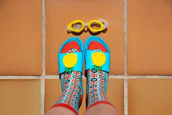 A young woman\'s feet in colorful socks and blue flip-flops next to yellow pool goggles. Summer concept