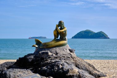 A mermaid statue, located on Samila Beach, sitting on a rock by the sea with a small island in the background clipart