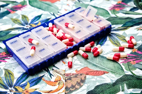 Days of the week pillbox full of red and white pills on top of a table with a tropical pattern.