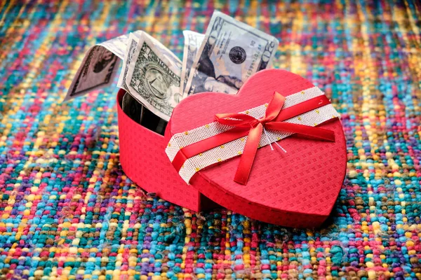Gift in the shape of an open heart and inside full of dollars, on a table with many colors. Happy Valentine\'s Day, Mother\'s Day, birthday gift.