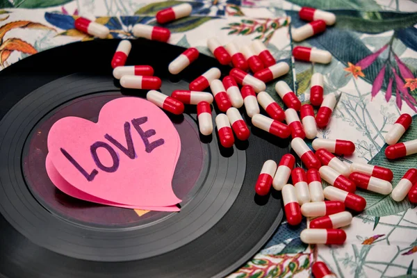 Vinyl record with a pink heart written the word love and a bunch of pills, on top of a table with a tropical pattern.