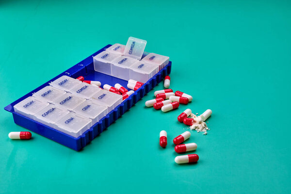 Pillbox of the days of the week on a green table with some red and white capsules on top. Photography in landscape format. Copy space.