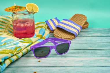 Şemsiyeli, bir dilim limonlu, mor güneş gözlüklü, havlulu ve plaj sandaletli, ahşap bir masa ve yeşil bir arka plan üzerinde ferahlatıcı pembe bir içecek..