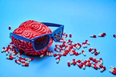 Mavi güneş gözlüklü, kırmızı bir beynin betimlemesi ve mavi bir yüzey üzerinde bir sürü hap..