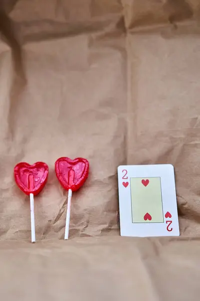 Two red heart-shaped lollipops and a two hearts card on a cardboard background.