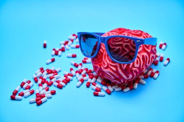 Mavi bir yüzeyde beyaz ve kırmızı kapsüllerle çevrili güneş gözlüklü kırmızı kauçuk beyin. Rahatlama kavramı.