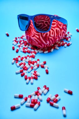 Mavi bir yüzeydeki kırmızı ve beyaz kapsüllerin tepesinde güneş gözlüklü kırmızı beyin..