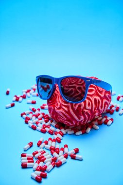 Kırmızı kauçuk beyin güneş gözlüğü takıyor ve mavi bir masada kırmızı ve beyaz haplarla çevrili. Stres kavramı.