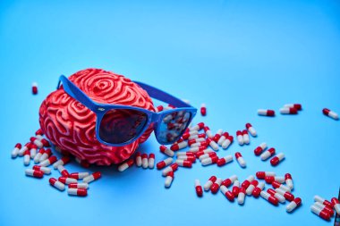 Kırmızı plastik insan beyni mavi güneş gözlüğü takıyor. Mavi arka planda kırmızı kapsüllerle çevrili. Sarhoşluk kavramı.