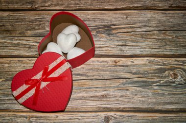 Kırsal bir arka planda, beyaz kalplerle dolu, açık kırmızı bir kalp şeklinde bir hediye kutusu yatıyor.