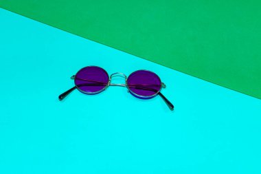 Mavi ve yeşil arka planda yuvarlak mor güneş gözlüğü.