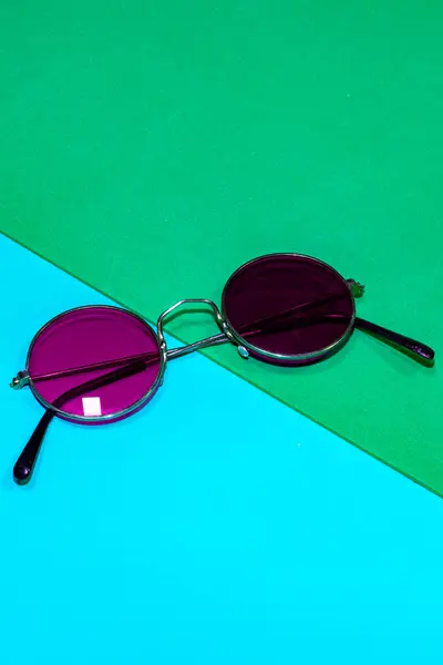 Mavi ve yeşil arka planda yuvarlak mor güneş gözlüğü.