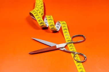 Keskin paslanmaz çelik makasın yanına yerleştirilmiş terzi elbisesi ölçüm bandının üst görüntüsü turuncu arka planda