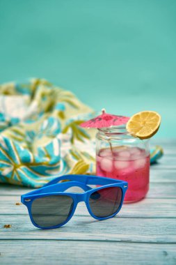 Bol buzlu, şemsiyeli, limonlu, mavi güneş gözlüklü, ahşap bir masa üzerinde tropik desenli plaj havlusuyla ferahlatıcı bir yaz içeceği..