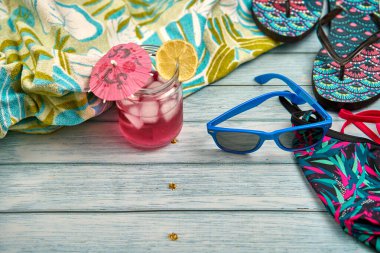 Karpuz ve limon suyu, mavi güneş gözlüğü, parmak arası terlik, bikini ve ahşap masa üzerinde tropik desenli bir plaj havlusu..
