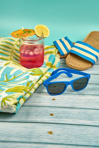 Şemsiyeli, limonlu, mavi güneş gözlüklü, havlulu, plaj sandaletli, ahşap bir masa ve yeşil bir arka planda ferahlatıcı pembe bir içecek..
