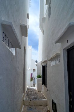 Beyaz binalardan oluşan dar bir ara sokak Menorca 'nın Binibeca köyünde gökyüzüne doğru gidiyor. Güneş, kaldırım taşı yolunda yumuşak bir ışık saçıyor..