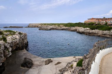 Binibeca, Menorca 'da kayalık kıyı şeridini ve berrak bir gökyüzünün altındaki sakin denizi gösteren manzaralı bir manzara. Fotoğraf, huzurlu ve resimli bir kıyı manzarası sunuyor..