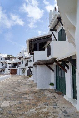Binibeca, Menorca 'daki beyaz boyalı binaların ve kaldırımlı sokakların mavi gökyüzünün altında pofuduk bulutların altında benzersiz bir mimariyi gözler önüne seren bir perspektif..
