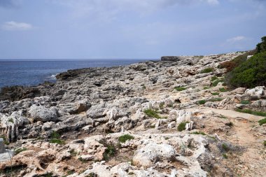 Binibeca, Menorca 'nın Rocky sahil şeridi bulutlu bir gökyüzünün altında sakin bir deniz. Manzarada çeşitli kaya oluşumları ve kıyı bitkileri yer almaktadır..