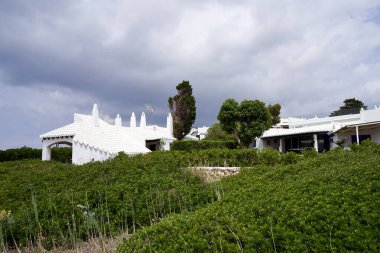 Binibeca, Menorca 'nın pitoresk manzarası, bulutlu bir gökyüzü altında yemyeşil bir zemin üzerine kurulmuş beyaz binalar..