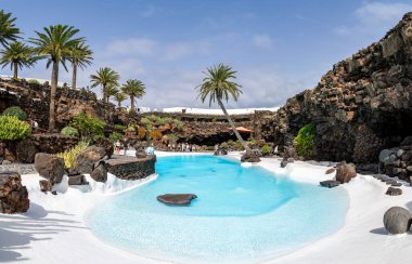 Lanzarote 'deki Jameos del Agua' nın göz kamaştırıcı manzarası, açık mavi gökyüzünün altında siyah volkanik kayalar ve yemyeşil palmiye ağaçlarıyla çevrili bir turkuaz havuzu içerir..