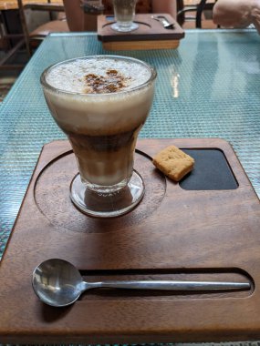 Küçük bir bisküvi ile ahşap bir tepside zarif bir şekilde servis edilen katmanlı bir kahve içeceği, bir anlık keyfi yakalar. Masa ayarı çok parlak..