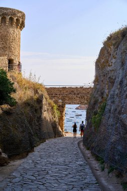 Tossa de Mar 'da taş duvarlar ve iki kişinin yürüdüğü tarihi bir kuleyle çevrili manzaralı bir patika denize açılıyor..