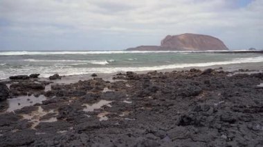 La Graciosas kayalık sahili bulutlu bir gökyüzü altında, dalgalar çarpıyor ve uçan bir kuş.