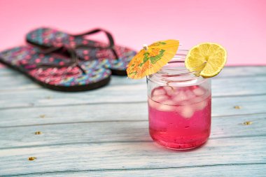 Şemsiyeli karpuz içeceği ve arka planda parmak arası terlik ve pembe bir arka plan olan ahşap bir masada bir dilim limon..