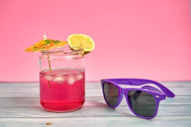 Şemsiyesi ve ahşap bir masa üzerinde bir dilim limon ve mor güneş gözlüğü ile ferahlatıcı pembe bir içecek ve pembe bir arka plan..