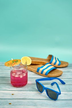 Şemsiyeli, limonlu, mavi güneş gözlüklü, plaj sandaletli, ahşap bir masa ve yeşil bir arka planda ferahlatıcı pembe bir içecek..