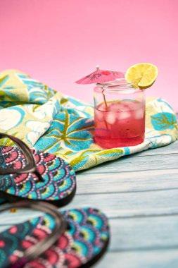 Şemsiyeli Sangria içeceği ve plaj havlusunda bir dilim limon tropikal desenli ve pembe arka planlı terlikler..