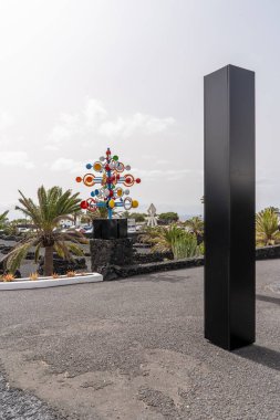 Siyah bir dikilitaşın yanında canlı renkli bir heykel duruyor. Bu sanat sahnesi İspanya 'nın Lanzarote şehrinde geçiyor..