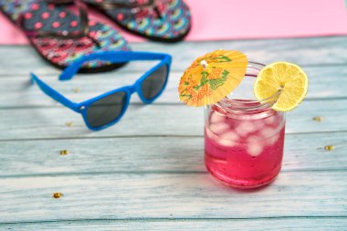 Şemsiyeli karpuz ve limon suyu ve bir dilim limon, mavi güneş gözlüğü ve pembe arka planlı ahşap bir masada parmak arası terlikler..