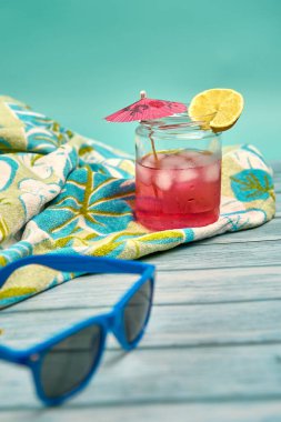 Tazelenmiş pembe bir şemsiye, bir dilim limon, mavi güneş gözlüğü ve ahşap bir masa üzerinde tropik desenli bir plaj havlusu ve mavi bir arka plan..