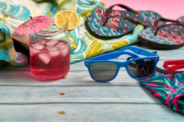 Karpuz ve limon suyu, mavi güneş gözlüğü, parmak arası terlik, bikini ve ahşap masa üzerinde tropik desenli bir plaj havlusu..