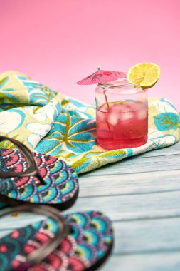Pembe bir içecek, şemsiye ve plaj havlusunda bir dilim limon tropikal desenli ve pembe arka planlı terlikler..
