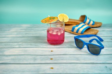 Şemsiyeli, limonlu, mavi güneş gözlüklü, plaj sandaletli, ahşap bir masa ve yeşil bir arka planda ferahlatıcı pembe bir içecek..