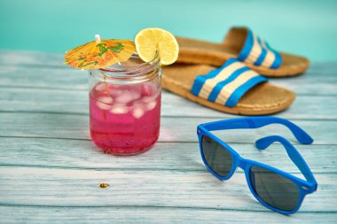 Şemsiyeli, limonlu, mavi güneş gözlüklü, plaj sandaletli, ahşap bir masa ve yeşil bir arka planda ferahlatıcı pembe bir içecek..