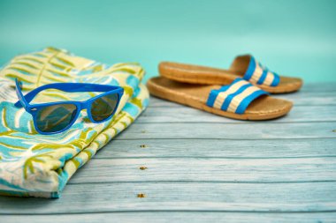 Mavi güneş gözlüğü, sandalet ve plaj havlusu, ahşap masa ve yeşil arka plan.