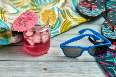 Sangria ve limon suyu, mavi güneş gözlükleri, parmak arası terlikler, bikini ve ahşap masa üzerinde tropik desenli bir plaj havlusu..