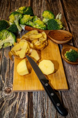 Keskin bir bıçak, ahşap bir kesme tahtasına serilir. Tahta bir yüzey üzerinde brokoli çiçekleriyle patates soyulur ve kesilir..