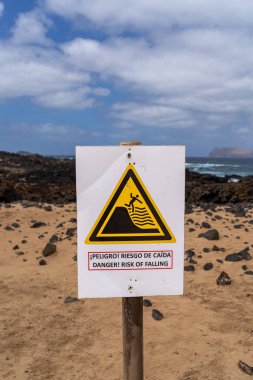 Düşme riskini gösteren bir uyarı işareti, arka planda mavi gökyüzü ve okyanus olan kumlu bir plaja asılır..