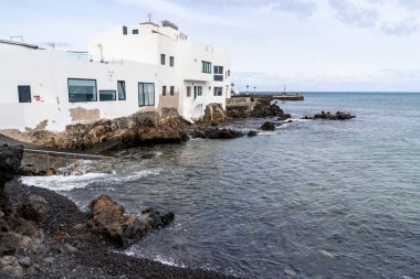A seaside building in Punta Mujeres, Lanzarote, with white walls and dark window frames. Rocky shoreline and calm sea meet under a cloudy sky. Lanzarote, Spain, 2025-07-20