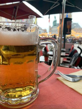 Açık bir kafede kırmızı bir masada, cam bardakta buz gibi bir bira. Arka planda binalar ve insanlarla dolu bir şehir caddesinin bulanık bir görüntüsü var..