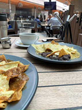  Açık hava bir kafede rahat bir yemek deneyiminin fotoğrafı. Siyah fasülyeli iki tabak chilaquiles, bir fincan kahveyle birlikte ahşap bir masada sergilenir.